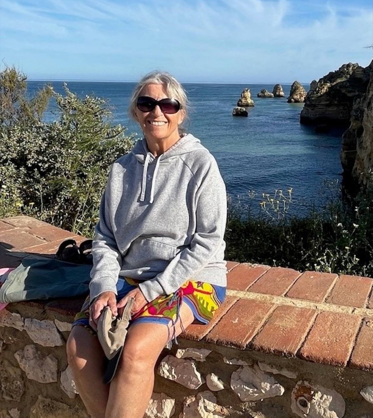 Jan Barrie sitting on a stone wall above a turquoise bay, smiling in sunglasses